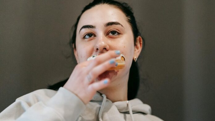 Glad young brunette in comfy hoodie biting delicious glazed doughnut and looking away in light room