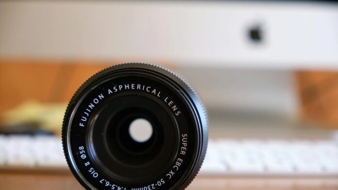 Close-up of a Fujinon aspherical lens with a blurred computer in the background, on a wooden desk.