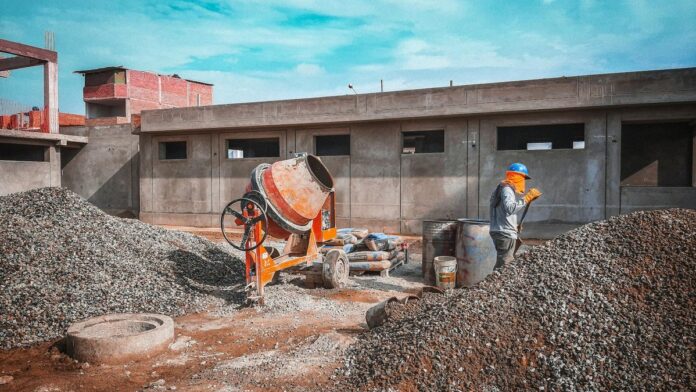 A construction site with a worker wearing safety gear next to a cement mixer and gravel piles.