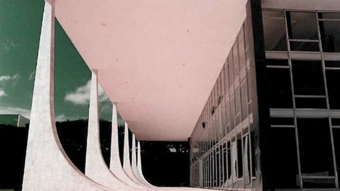 Silvio Almeida de terno e gravata, em plano médio, olhando seriamente em direção à câmera. Fundo neutro de escritório.