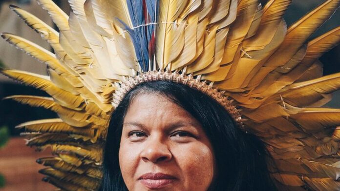 Foto de Sonia Guajajara, vestindo roupas e colares indígenas, em entrevista com expressão séria.