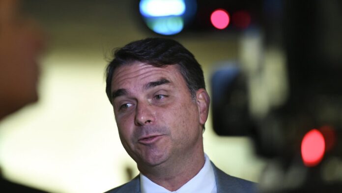 Foto de Flávio Bolsonaro discursando ao microfone em plenário, com expressão séria e gestos das mãos.