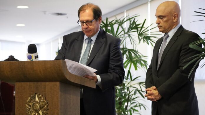 Cláudio Castro em pé ao lado de outros homens em um plenário do tribunal, com bandeira do Brasil ao fundo.
