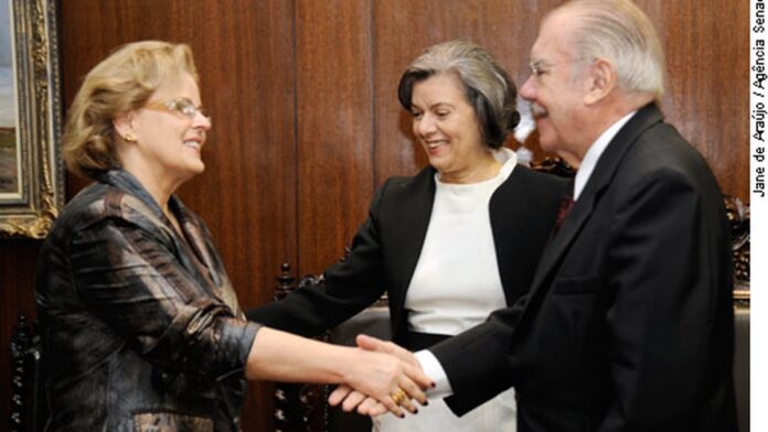 Mãos de uma mulher grávida repousam sobre seu ventre em um ambiente de escritório com documentos ao fundo.