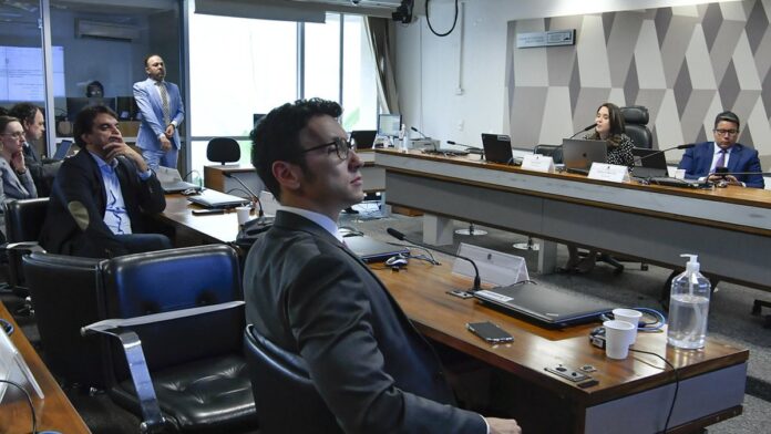 Homem de terno fala em um microfone diante de uma plateia em um auditório, com uma tela ao fundo.