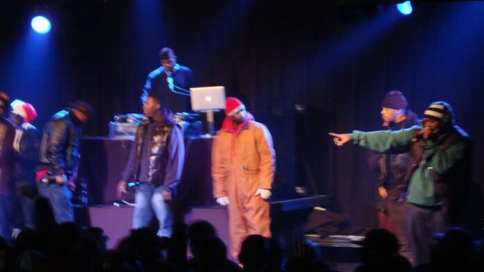 Membros do Wu-Tang Clan se apresentam em palco com iluminação azul e microfones em mãos.