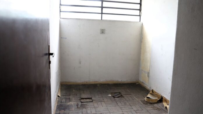 Interior da cela do DOI-CODI, com paredes de alvenaria desgastadas e marcas de desgaste que remetem ao período militar.