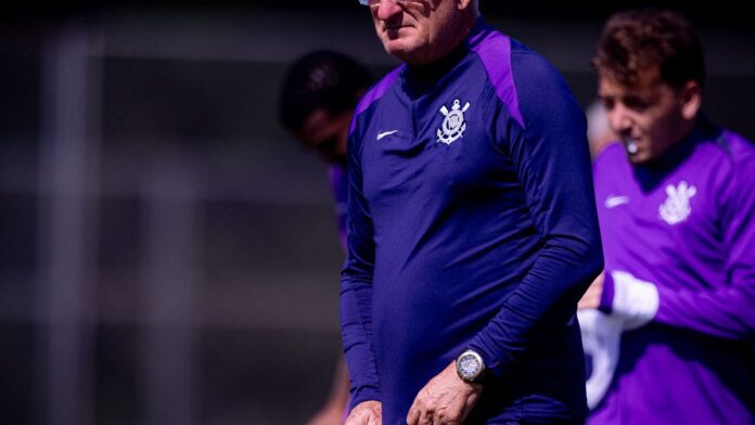Dorival Júnior com semblante sério, vestindo agasalho esportivo, em frente a uma placa com o escudo do Corinthians.