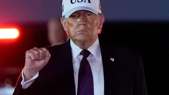 Donald Trump gesticula em discurso, com expressão séria, em cenário com bandeiras dos EUA ao fundo.