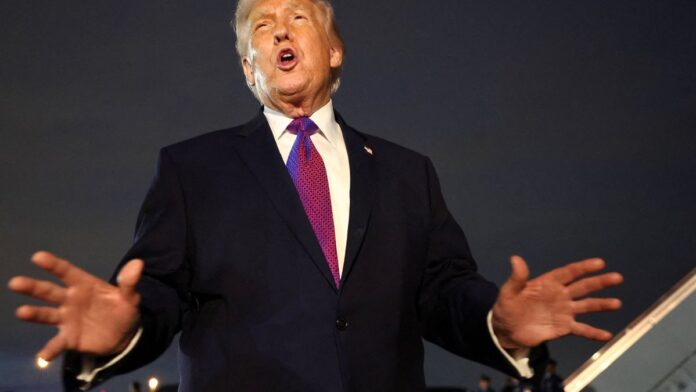 Donald Trump em um palanque, gesticulando e discursando com expressão séria diante de bandeiras americanas.
