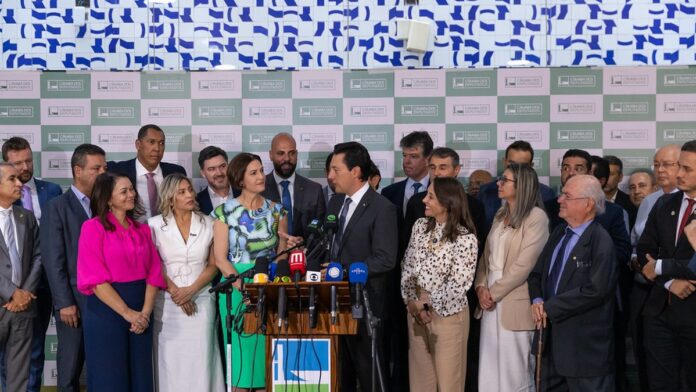 Cristina Graeml sorri ao assinar documentos de filiação partidária, cercada por lideranças políticas em uma sala.