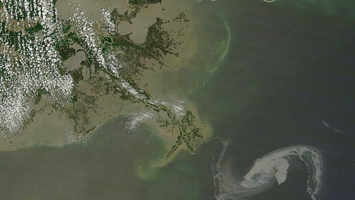 Plataforma de petróleo operando no mar, com grande mancha escura na superfície da água e céu nublado ao fundo.