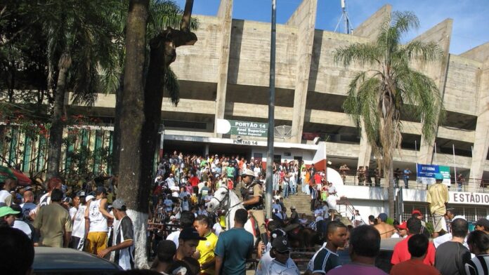 Jogadores de futebol em campo de gramado durante partida do clássico Gre-Nal com torcida ao fundo nas arquibancadas.