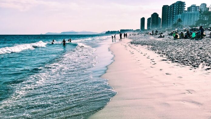 helic-ptero-cai-na-praia-da-barra-da-tijuca-o-que- Helicóptero caído sobre a areia da praia, cercado por equipes de resgate e curiosos sob céu claro.