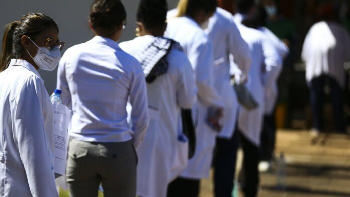 Médico de jaleco branco atende paciente em consultório moderno, com estetoscópio e prontuário sobre a mesa.