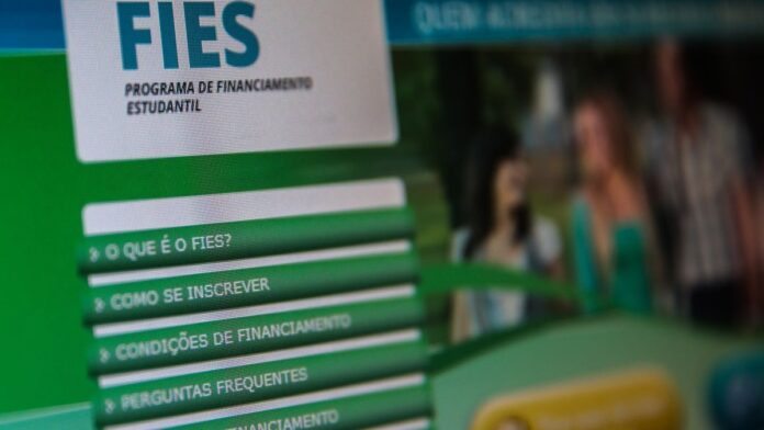 Estudante sentado em frente a um notebook, concentrado, com papéis e materiais de estudo sobre uma mesa.