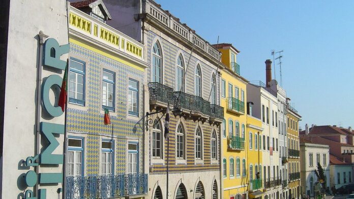 Pessoas em fila em frente a um edifício público de arquitetura clássica em Portugal, com bandeiras hasteadas ao fundo.