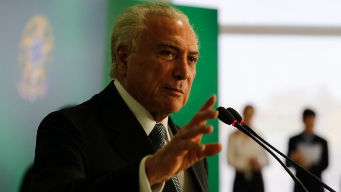 Pessoa pilotando uma motocicleta em uma via urbana, vestindo apenas calça, capacete e luvas.