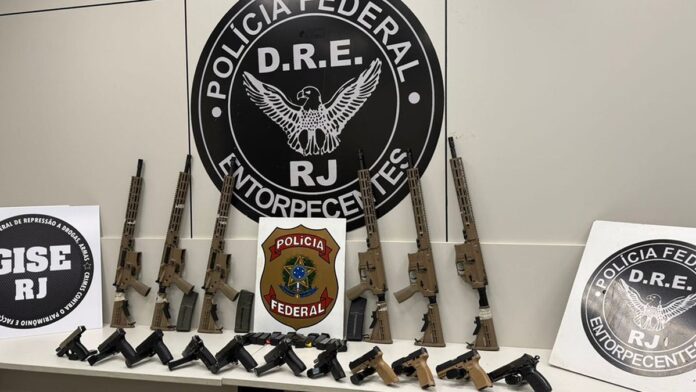 Armamento apreendido pela polícia, incluindo diversos fuzis e pistolas, disposto sobre uma mesa em fundo neutro.