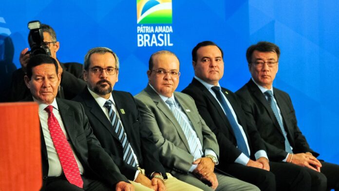 Renato Bolsonaro posa sorridente, vestindo camisa polo azul, segurando cartaz com o número 2222 em evento partidário.