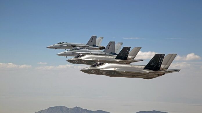 resgate-no-ir-for-as-dos-eua-buscam-segundo-piloto Equipe militar em helicóptero sobrevoa região montanhosa e árida em busca de aeronave acidentada no horizonte.