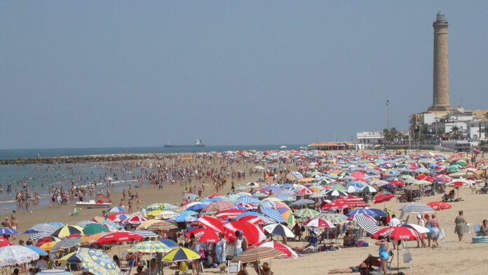 Ondas gigantes avançam em direção a uma costa urbana espanhola, sob um céu nublado e ameaçador.
