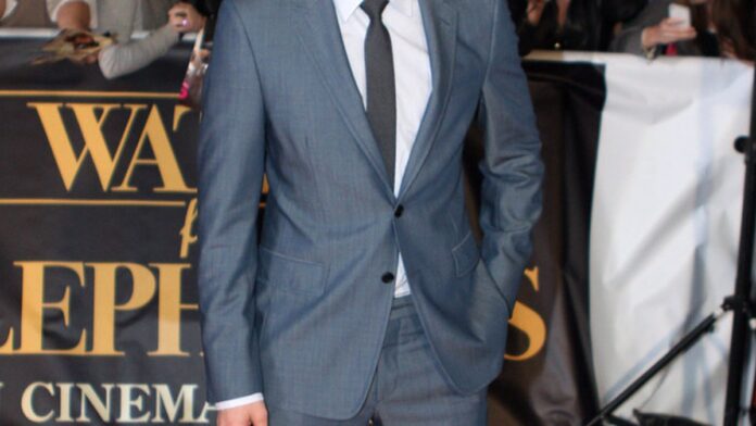 Robert Pattinson posa segurando uma garrafa de cerveja 1664 Blanc, vestindo um terno elegante em ambiente sofisticado.