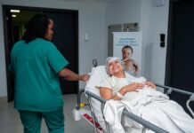 Hospital Oncológico Infantil em Belém promove oficina de culinária para pacientes A caring nurse assists a patient in a hospital corridor with a joyful demeanor.