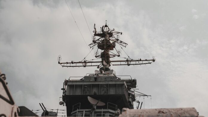 Close-up view of a military warship's radar and antenna mast against a cloudy sky.