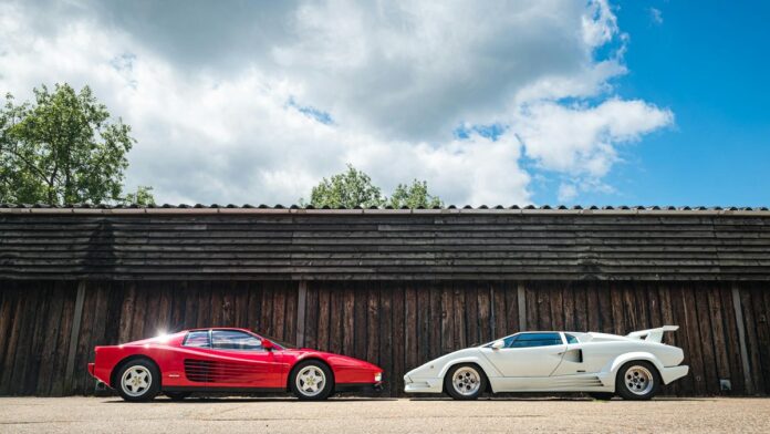 Red Ferrari Testarossa and white Lamborghini Countach parked outdoors against wooden background.