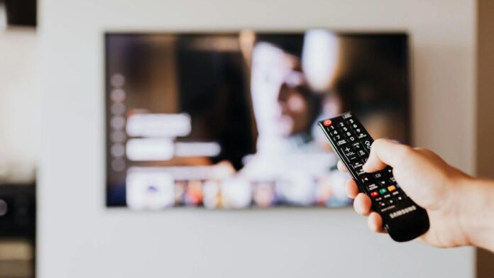 Person using a TV remote control with a blurred television screen in the background.