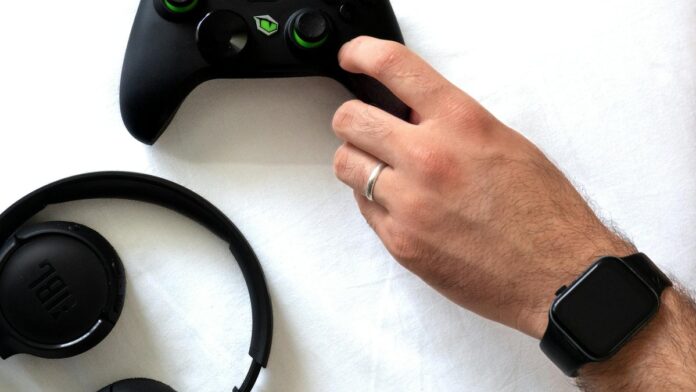 Hand holding a gaming controller with headphones and smartwatch on white background.