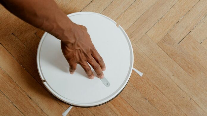 A close-up of a hand activating a robotic vacuum cleaner on a wooden floor.