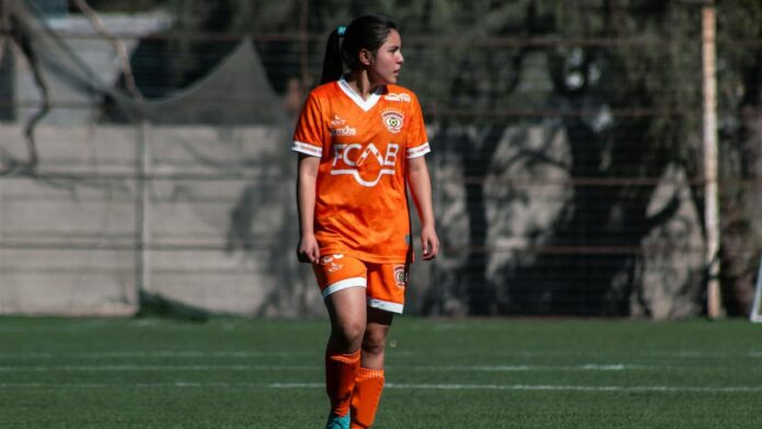 A female soccer player in orange uniform during a match on an outdoor field in Chile.