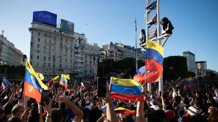 A large crowd waves Venezuelan flags during a protest in a bustling city square.