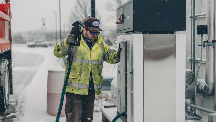 s4-6fc4ef495a1c A man wearing a jacket refuels a truck at a gas station during winter with snow falling.