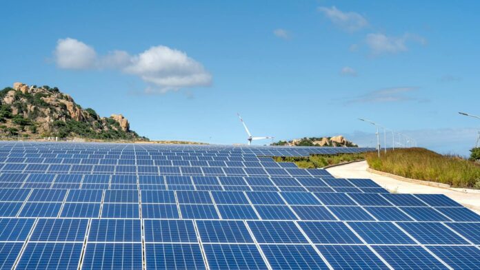 Expansive solar farm with rows of panels under clear blue sky and hills in the background.