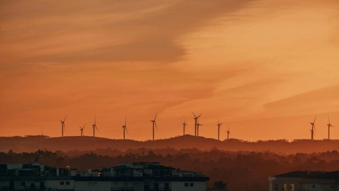 s4-8188ccc9a426-17 A picturesque view of wind turbines against an evening sunset sky over a silhouetted urban landscape.