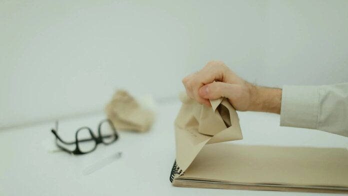 A close-up of a hand crumpling paper on a desk with glasses in the background, evoking frustration.