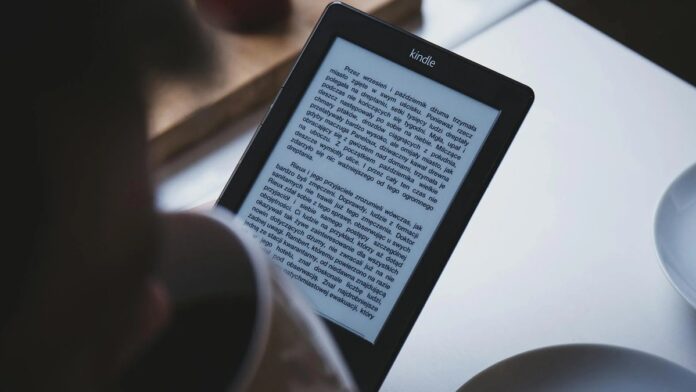 A person reads on a Kindle e-reader while holding a cup of coffee, indoors.