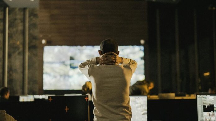 A man stands thoughtfully in a dimly lit control room with various screens.