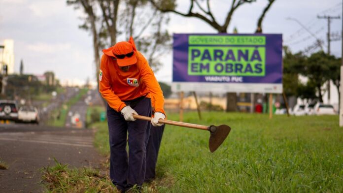 A worker wearing orange uniform maintains roadside grass in Londrina, Brazil.