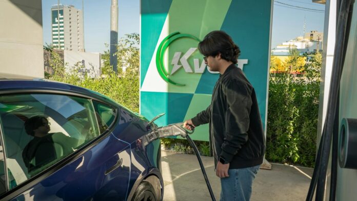 Young man charging a Tesla Model 3 at an outdoor electric vehicle station during the day.