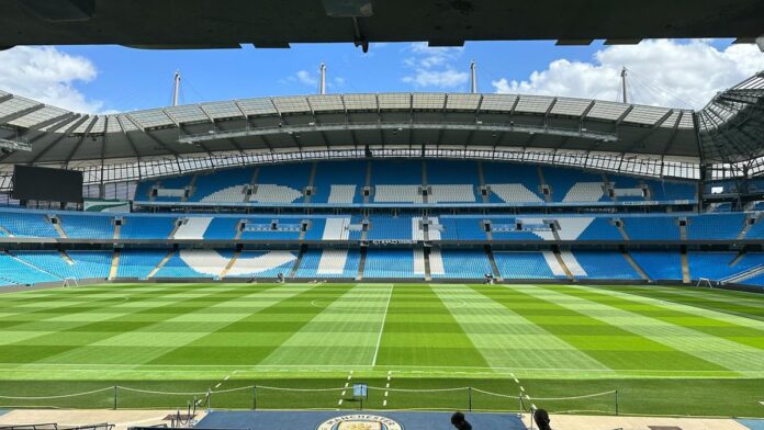 s4-eb80b301dce4-1 Panoramic view of Etihad Stadium, showcasing the empty football pitch and stands under a clear blue sky.
