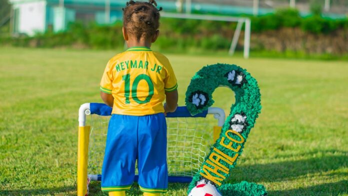 A young child wearing a Neymar Jr. jersey stands by a mini soccer goal on a grassy field.