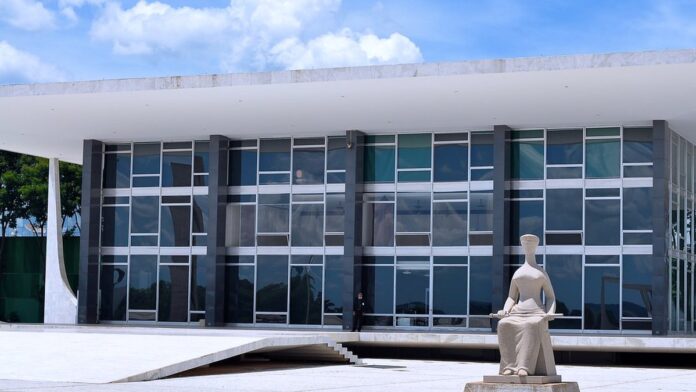 Fachada de um fórum de justiça com colunas de concreto e bandeiras do Brasil ao fundo, sob luz do dia.